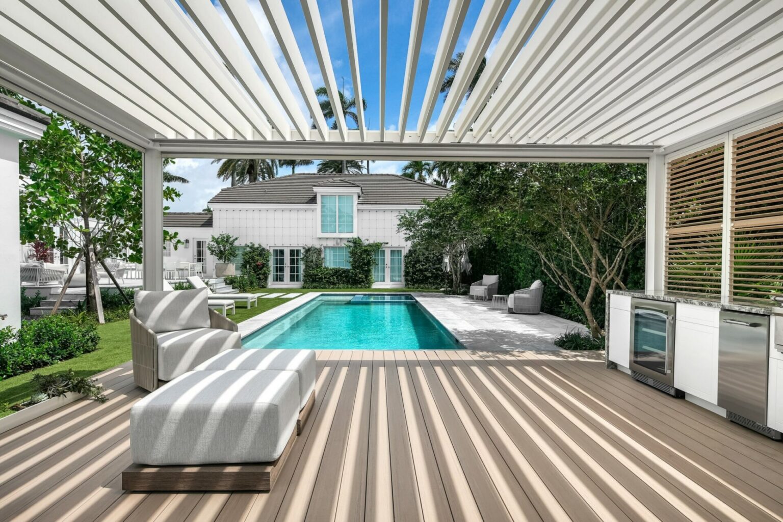 Modern outdoor patio with striped shadow patterns, lounge chairs, and a built-in kitchenette overlooks a rectangular pool surrounded by greenery and a white house with blue windows under a clear sky.